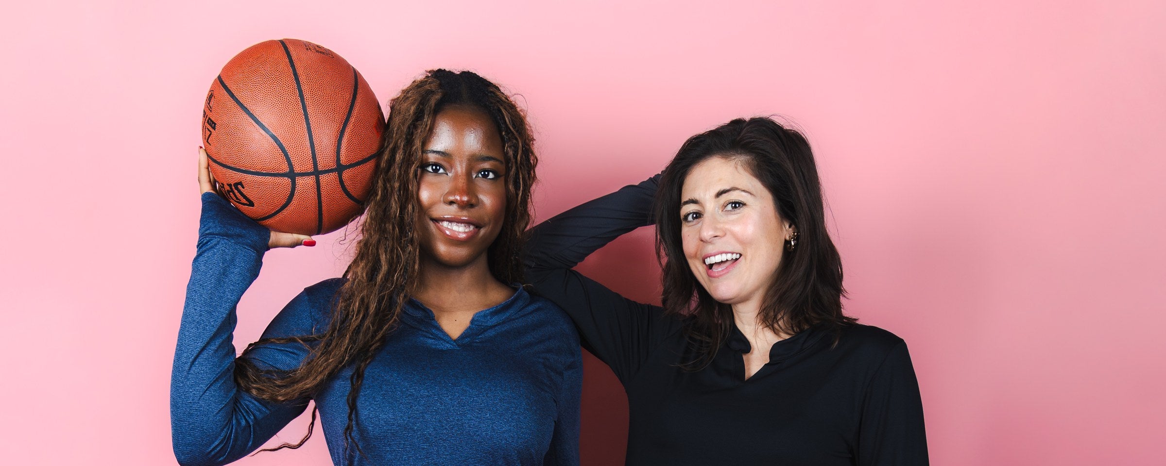 Two women in modest activewear holding a basketball and a soccer ball against a pink studio backdrop